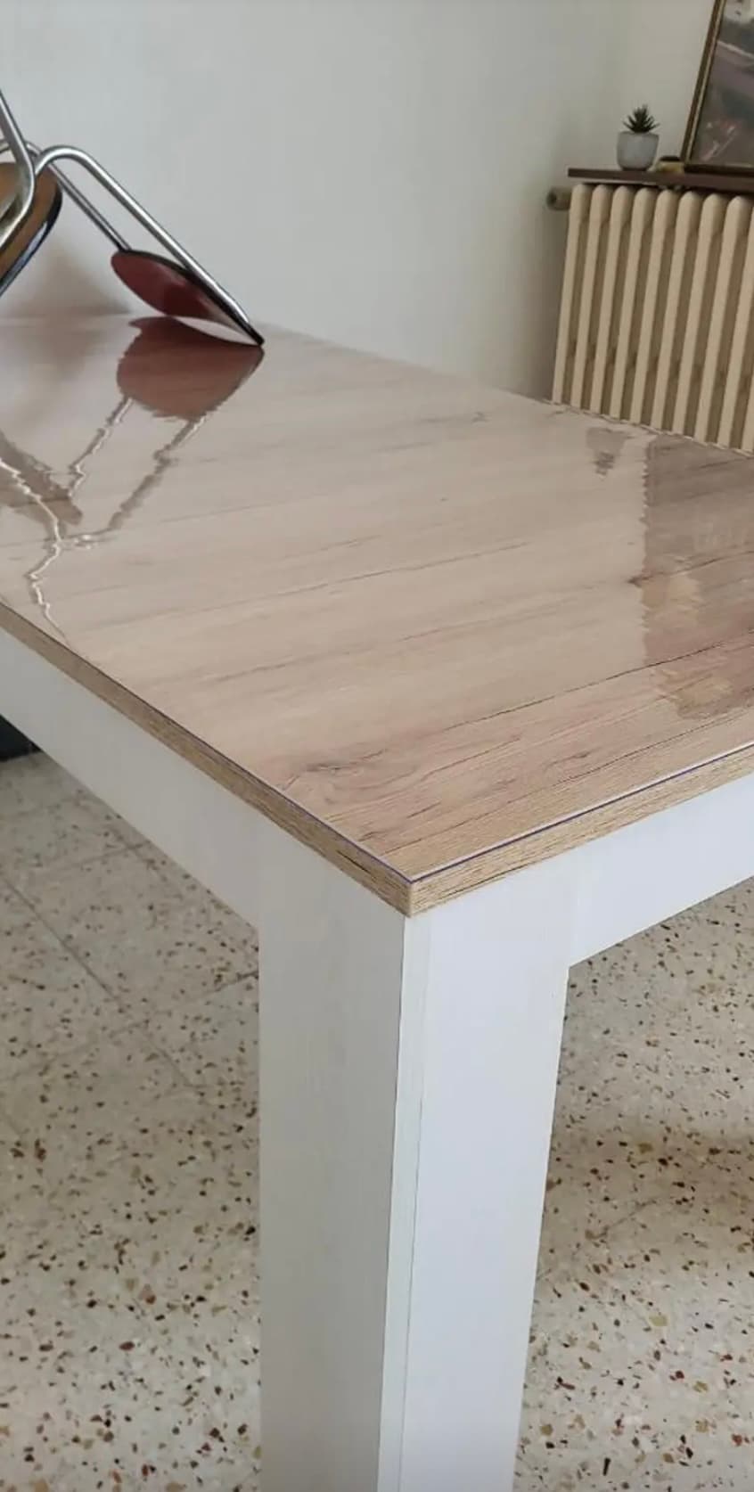 Svetlá stolová doska na bielych nohách s matným leskom; liatinový radiátor v pozadí