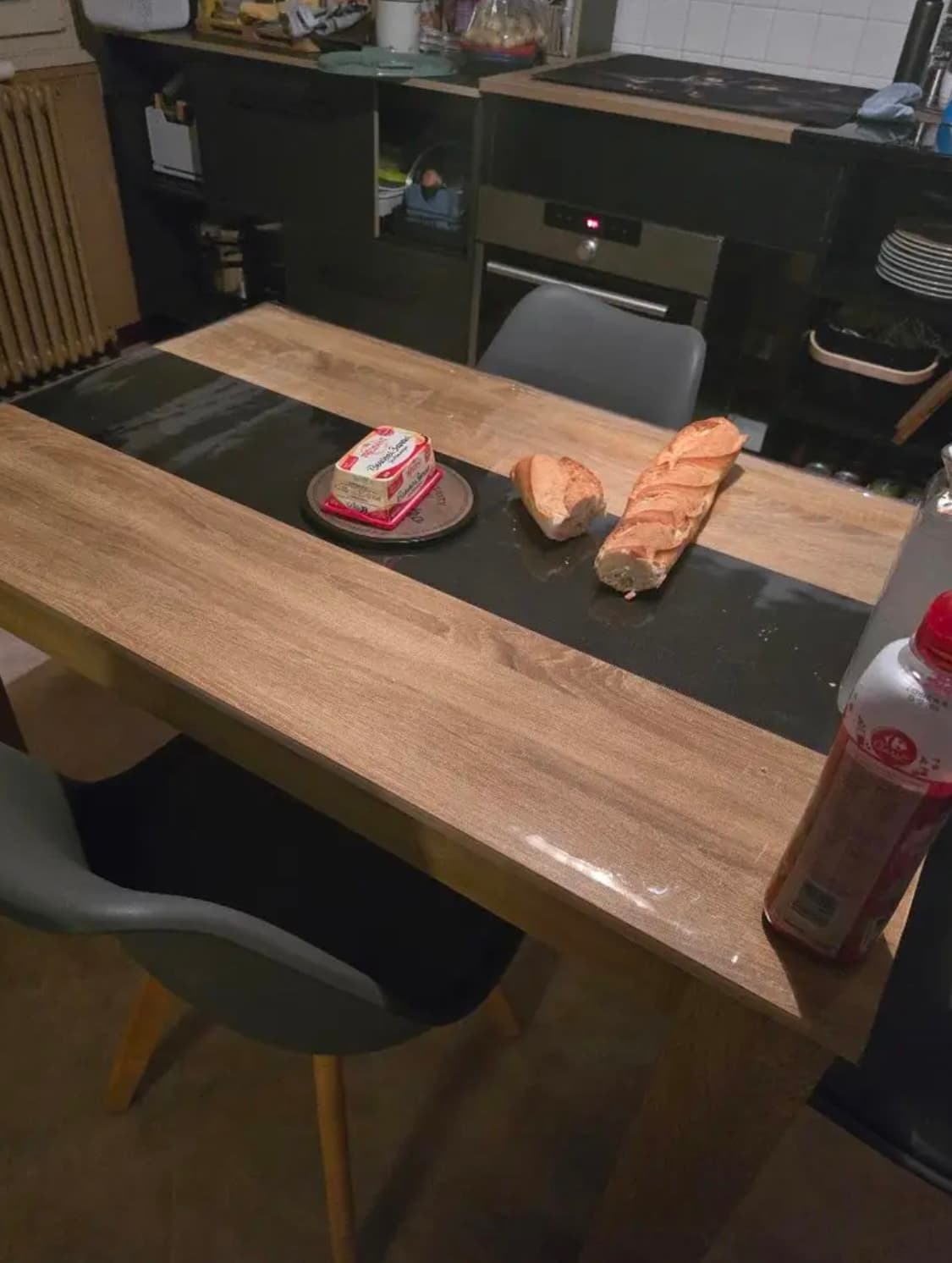 Kuchynský stôl s tmavým pruhom v strede, na ňom bageta a maslo; stolová doska pod mäkkým sklom