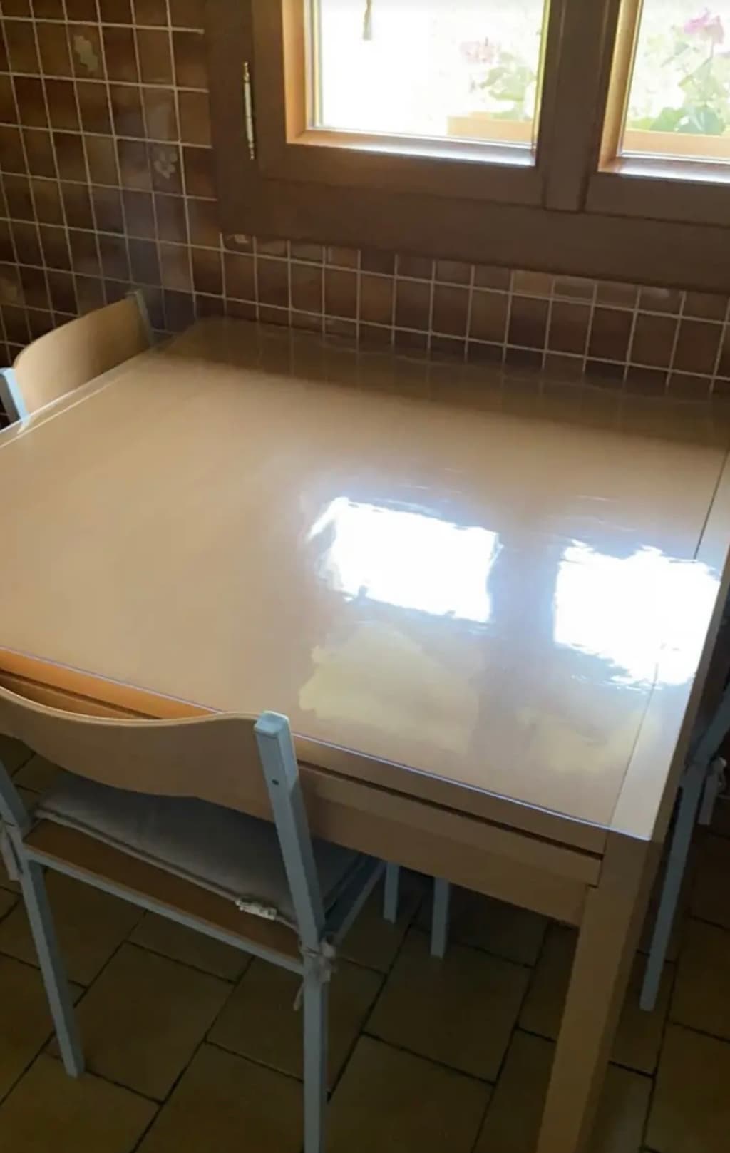 Malý krémový stôl v kuchyni s dlaždicami; na pulte je podložka a v okne črepníky s muškátmi
